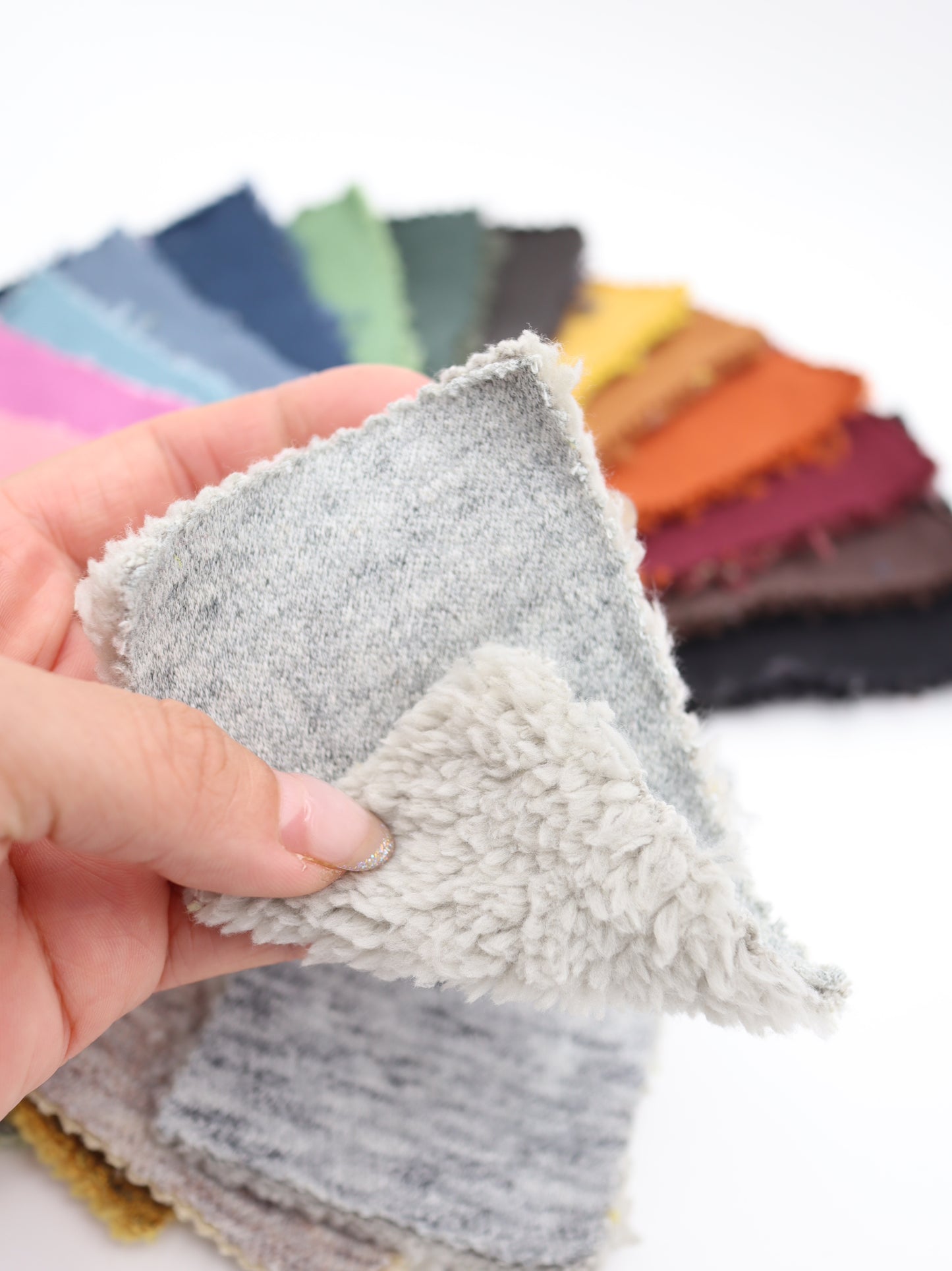 リバーシブルWフェイス裏ソフトボアスウェット生地の接写画像。裏面は柔らかい肌触りのボア、表面は杢調のニットになっており、背景には複数の色の生地見本が並んでいる。